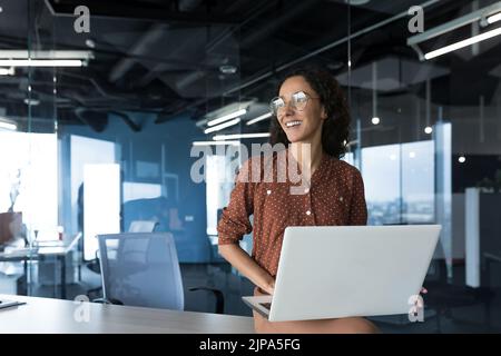 Portrait young beautiful woman with curly hair, successful woman in glasses smiling and looking out window, business woman freelancer working in office standing with laptop smiling looking out window Stock Photo