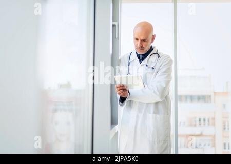 Portrait of careoworn male doctor holding a digital tablet in his hand while standing on hospital’s foyer. Stock Photo