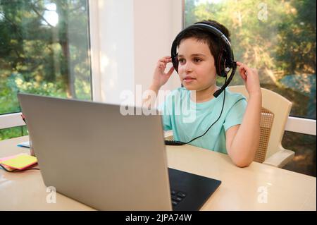 Handsome schoolboy in headphones using laptop watches broadcasted video lessons while studying remotely in online school Stock Photo