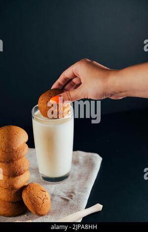 Oatmeal cookies laid out in a stack, a woman's hand soaks cookies in a milk glass. Black background. Food close-up Stock Photo