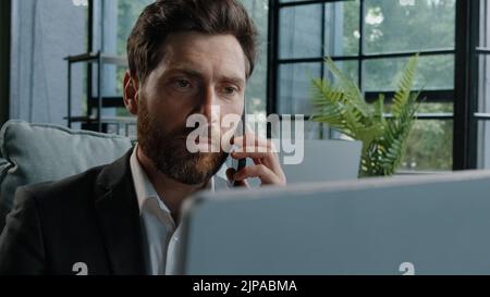 Angry bearded Caucasian adult 40s mad frustrated businessman arguing yelling conflict quarrel talking on phone looking at laptop computer sitting at Stock Photo