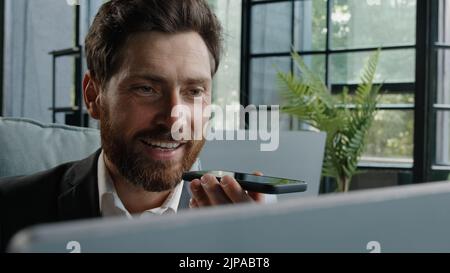 Happy adult 40s male manager bearded businessman sitting at workplace with computer smiling recording audio message sending voicemail using assistant Stock Photo