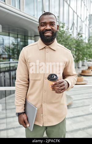 Vertical waist up portrait of smiling black man holding laptop and coffee in city Stock Photo
