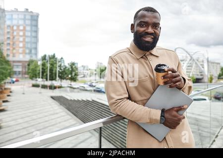 Waist up portrait of bearded black man holding laptop and coffee in city setting, copy space Stock Photo