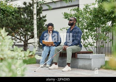 Full length portrait of adult couple with partner with visual disability sitting on bench in park together Stock Photo