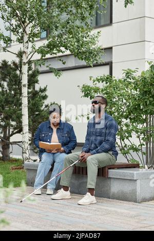 Vertical full length portrait of adult couple with partner with visual disability sitting on bench in park together Stock Photo