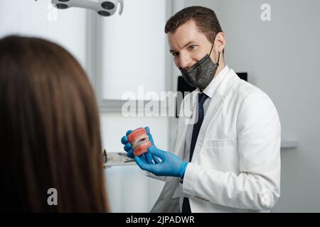 Male dentist holding tooth model with metal braces and speaks to his patient Stock Photo