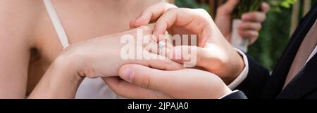 Cropped view of groom wearing ring on finger of bride outdoors, banner Stock Photo