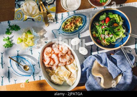 summer Seafood supper table spread to make fish tacos, prawns, guacamole, lime, tortilla, salsa on blue and white fish tablecloth in the sun Stock Photo