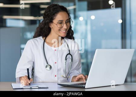 Young beautiful arab woman works in modern clinic office, doctor conducts online consultation with patients, worker at laptop in white coat and stethoscope, writes prescription Stock Photo