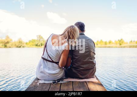 Senior family couple relaxing by summer lake. Man and woman enjoying natural landscape and hugging sitting on pier Stock Photo