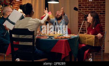 Diverse joyful people gathered in living room to celebrate winter holiday while giving each other presents. Festive relatives sitting at Christmas dinner table while exchanging gifts. Stock Photo