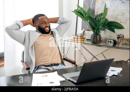 Happy calm african american man, freelancer, company employee, sits at the workplace in the office, takes a break from work, puts his hands behind his head, looks to the side, dreams of rest, smiles Stock Photo