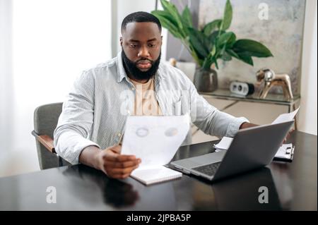Serious concentrated successful african american busy man, freelancer or company ceo, working with documents, sit at desk in modern office, working on a project uses laptop, studying financial graphs Stock Photo