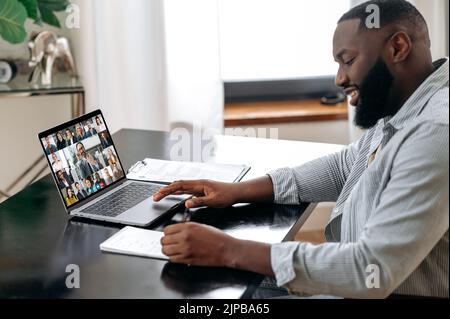 Online video briefing, seminar. African american man, sit at a desk in the office or at home, talking on a video conference with colleagues, listening to a lecture by a financial mentor, taking notes Stock Photo