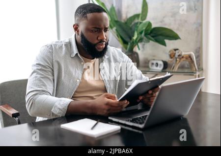 Serious focused clever african american busy business man, company ceo, working with documents, sit at desk in modern office, working on a project uses laptop, concentrated studying financial graphs Stock Photo