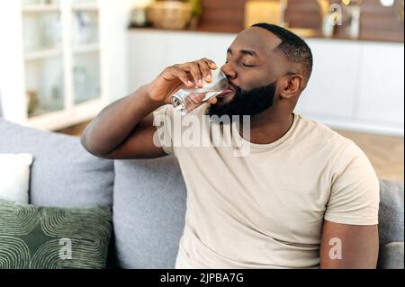 Healthy lifestyle. Positive African American guy, sit on sofa at home, drinking a glass of clean water. Handsome black man leads healthy lifestyle, monitors the balance of water in the body Stock Photo