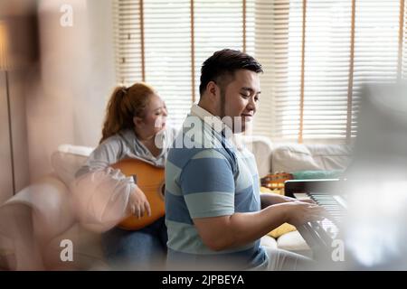 Young Asian chubby couple singing and playing acoustic guitar and piano together. Man and woman enjoying musical instrument. People in a band practicing in the house Stock Photo