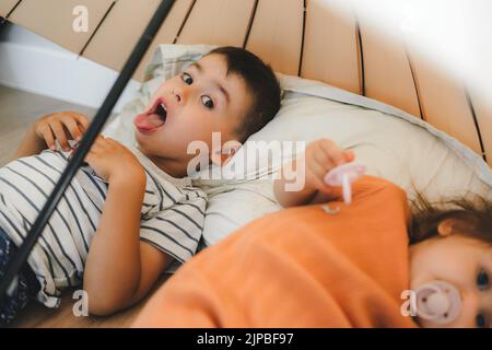 Boy showing his tongue while playing under the umbrella with his younger sister, in the comfortable house. Childhood concept. Stock Photo