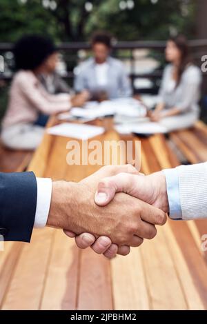 Lets shake on it. two unrecognizable businessmen shaking hands while their colleagues sit behind them outdoors. Stock Photo