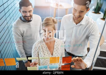 Theyre making progress. three young businesspeople brainstorming with sticky notes on a glass wall in the office. Stock Photo