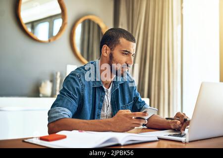I cant afford to loose my focus. a focused young man working on his laptop and checking his phone while being seated at a table at home. Stock Photo