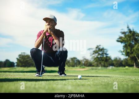 On the fairway. a focused young male golfer looking at a golf ball while being seated on the grass outside during the day. Stock Photo
