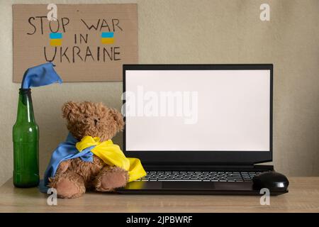 Teddy bear with the flag of Ukraine and a Molotov cocktail near a laptop with a white screen on the table, war in Ukraine, peace in Ukraine, a screen Stock Photo