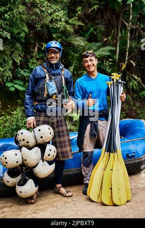 We got all the gear well ever need. Portrait of two cheerful men standing together next to a rubber boat while holding river rafting equipment outside Stock Photo