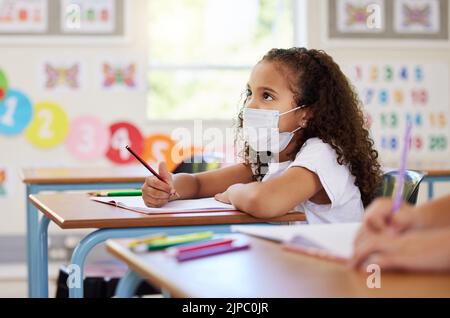 Education, classroom and learning with covid face mask on girl doing school work, writing and reading at her desk in elementary class. Elementary Stock Photo