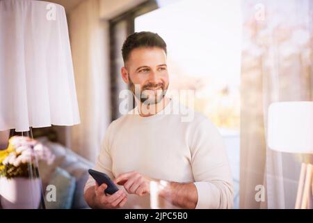 Shot of handsome man wearing casual clothes and text messaging while standing at the window at home. Stock Photo