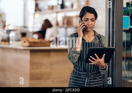 Female cafe business owner using phone talking, ordering and reading on tablet in her store. Serious businesswoman, entrepreneur or employee standing Stock Photo