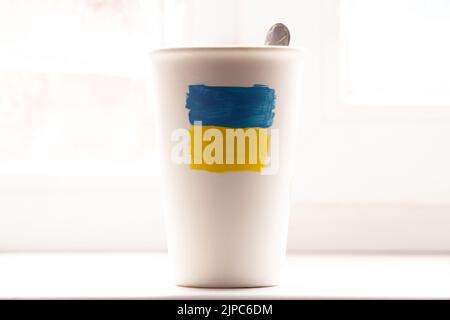 The flag of Ukraine yellow-blue is drawn on a white cup with coffee on a white table near the window close-up Stock Photo