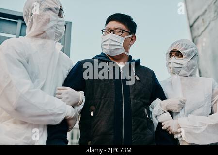 Asian man breaking covid regulation getting taken away or arrested by healthcare workers wearing hazmat protective suits. Male removed for not Stock Photo