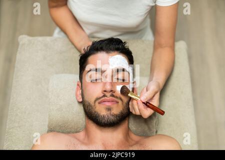 Top view of anonymous woman using soft brush to smear moisturizing mask on face of bearded man with closed eyes during spa session in salon Stock Photo