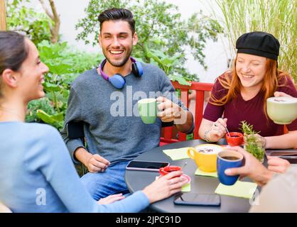 High angle of cheerful young men and women drinking coffee and laughing at joke while sitting around table during coffee break in garden Stock Photo