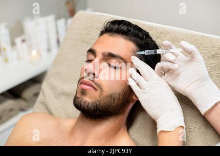 From above anonymous beautician in latex gloves injecting botulinum toxin into forehead of bearded young male client during cosmetology session in bea Stock Photo