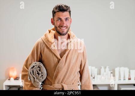 Cheerful young male client in warm bathrobe with rolled up towel looking at camera with smile against gray wall before massage in spa salon Stock Photo