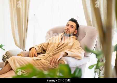 Bearded young male client with soft bathrobe looking at camera while chilling in comfortable armchair during spa session in beauty salon Stock Photo