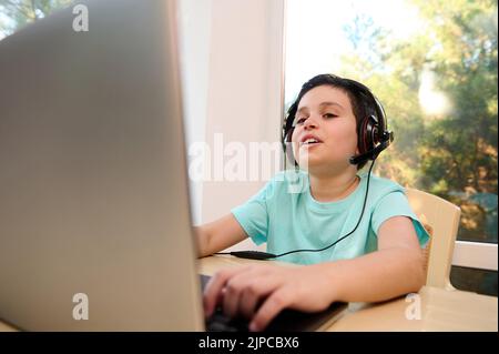 Spoiled, funny schoolboy in wireless headphones, sitting at laptop and watching an online video lesson. Back to school. Stock Photo