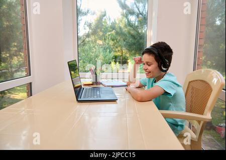 Handsome smart school boy, studying from home, sits at laptop and communicates with the teacher via video link. Stock Photo