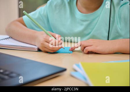 Details: hands of a school child holding a pencil and making notes during an online lesson on the laptop. Homeschooling Stock Photo
