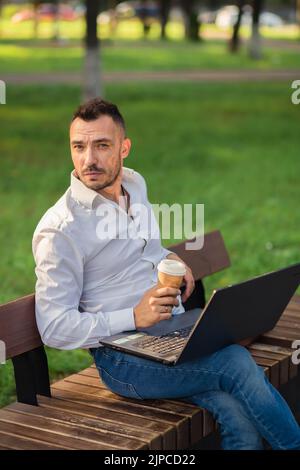 Man looking at the camera in the park with a laptop, drinking coffee. A young man on a background of green trees, a hot sunny summer day. Warm soft light, close-up. Stock Photo