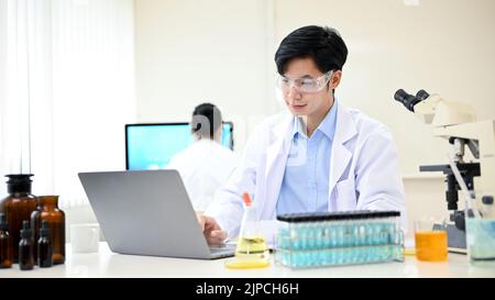 Intelligent and handsome young Asian male scientist or medical researcher working in his office, using laptop computer to check the chemical testing r Stock Photo