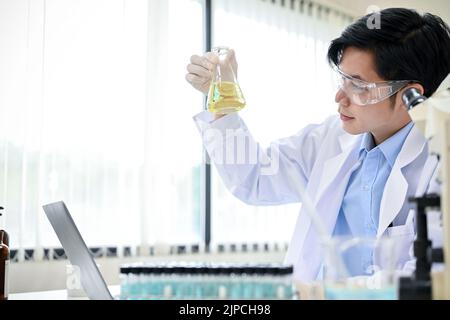 Handsome and smart young Asian male scientist or chemist in goggles and gown holding a laboratory flask, adjusting a liquid sample in a conical beaker Stock Photo