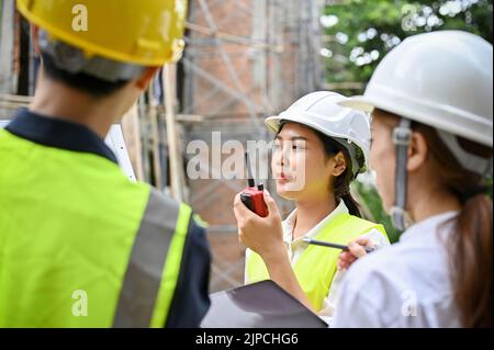 A professional young Asian female engineer using a walkie talkie to communicate with her worker in the construction site. Stock Photo
