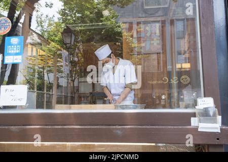 A shot through a window of a soba chef at Zenko-Ji Temple in summer wearing a mask and hat and preparing handmade buckwheat noodles for lunch. Stock Photo