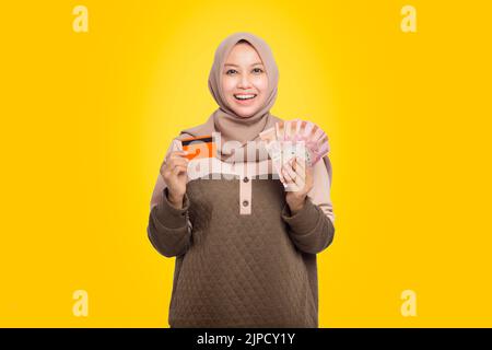 Portrait of happy Asian Muslim woman holding rupiah money and showing credit card isolated over yellow background. Indonesian Rupiah Stock Photo