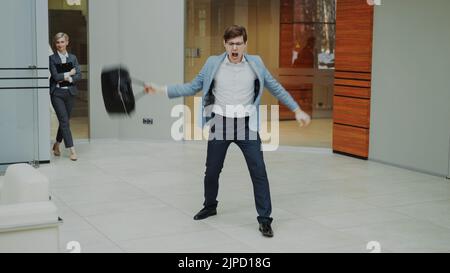 Crazy businessman dancing with briefcase in modern lobby indoors while his colleagues walking and watching him surprised Stock Photo