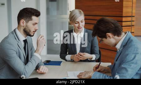 Young businessman in suit signing contract with his colleague while corporate lawyer explaining details of partnership sitting in modern office indoors Stock Photo
